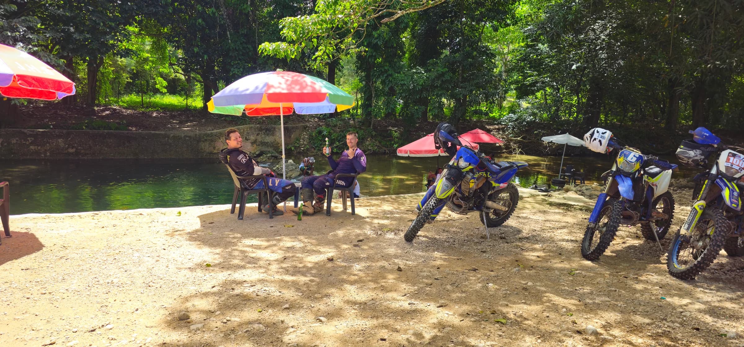 Riders resting under beach umbrellas at a river beach, bikes parked on the sand